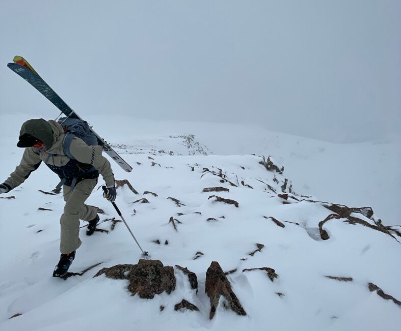 backcountry skier hiking in rocky snowy terrain with skis on his back