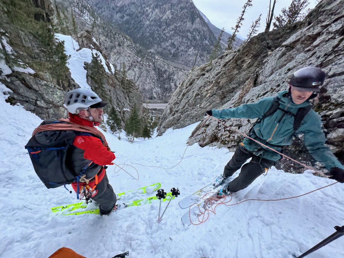 two skiers standing at the top a tight couloir wearing helmets