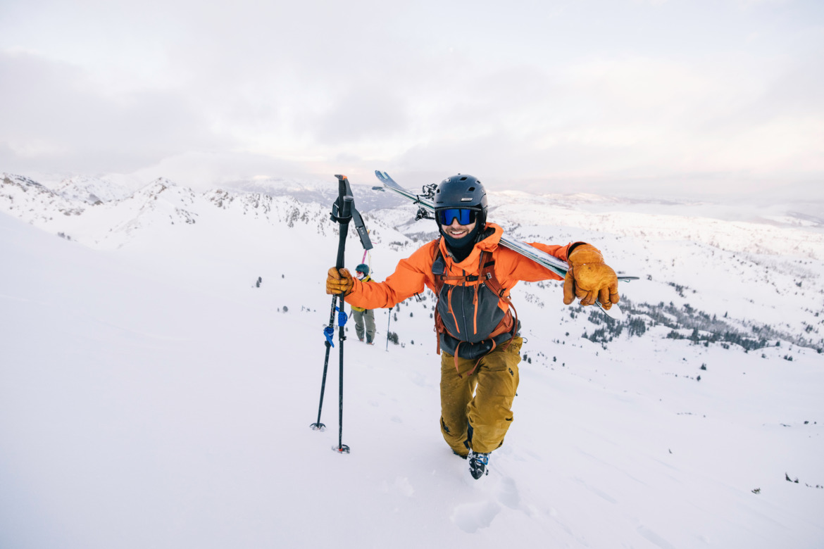 skier hiking with skis over his shoulder in orange jacket